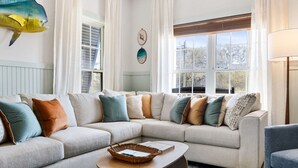 A living room with a beige sectional sofa, a round wooden coffee table, and a large window with curtains. - Folly - Large Front Porches - One Block to Beach (Folly Beach)