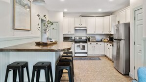 A modern kitchen with white cabinets, a stainless steel refrigerator, a built-in oven, and a bar area with stools. - Folly - Large Front Porches - One Block to Beach (Folly Beach)