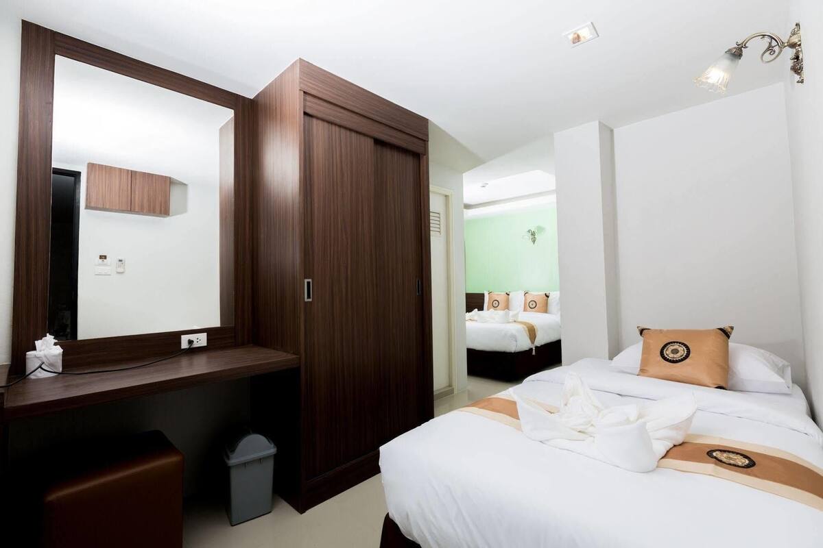 Habitación de hotel con armario de madera, un escritorio con cesto de basura, cama con ropa blanca y almohadones decorativos, y una pared de acento verde.