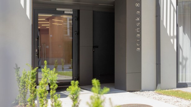 Una entrada de edificio moderna con puerta de cristal, un pequeño jardín con plantas y un cartel con el nombre "arminia 83".