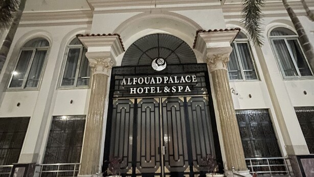 La entrada al Hotel & Spa Al Fouad Palace, con un imponente arco y un letrero, flanqueada por columnas y palmeras.