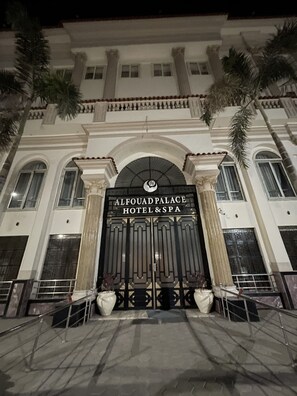 La entrada al Hotel & Spa Al Fouad Palace, con un imponente arco y un letrero, flanqueada por columnas y palmeras.