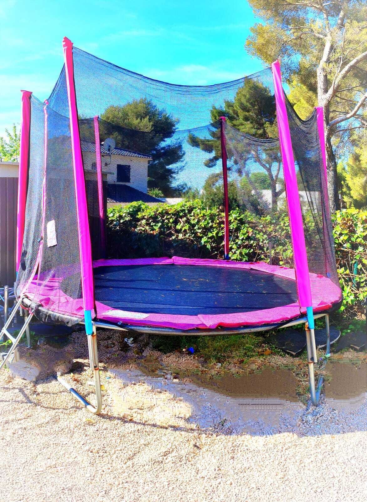 A trampoline with pink and blue safety netting, set up outdoors with a gravel ground and trees in the background.