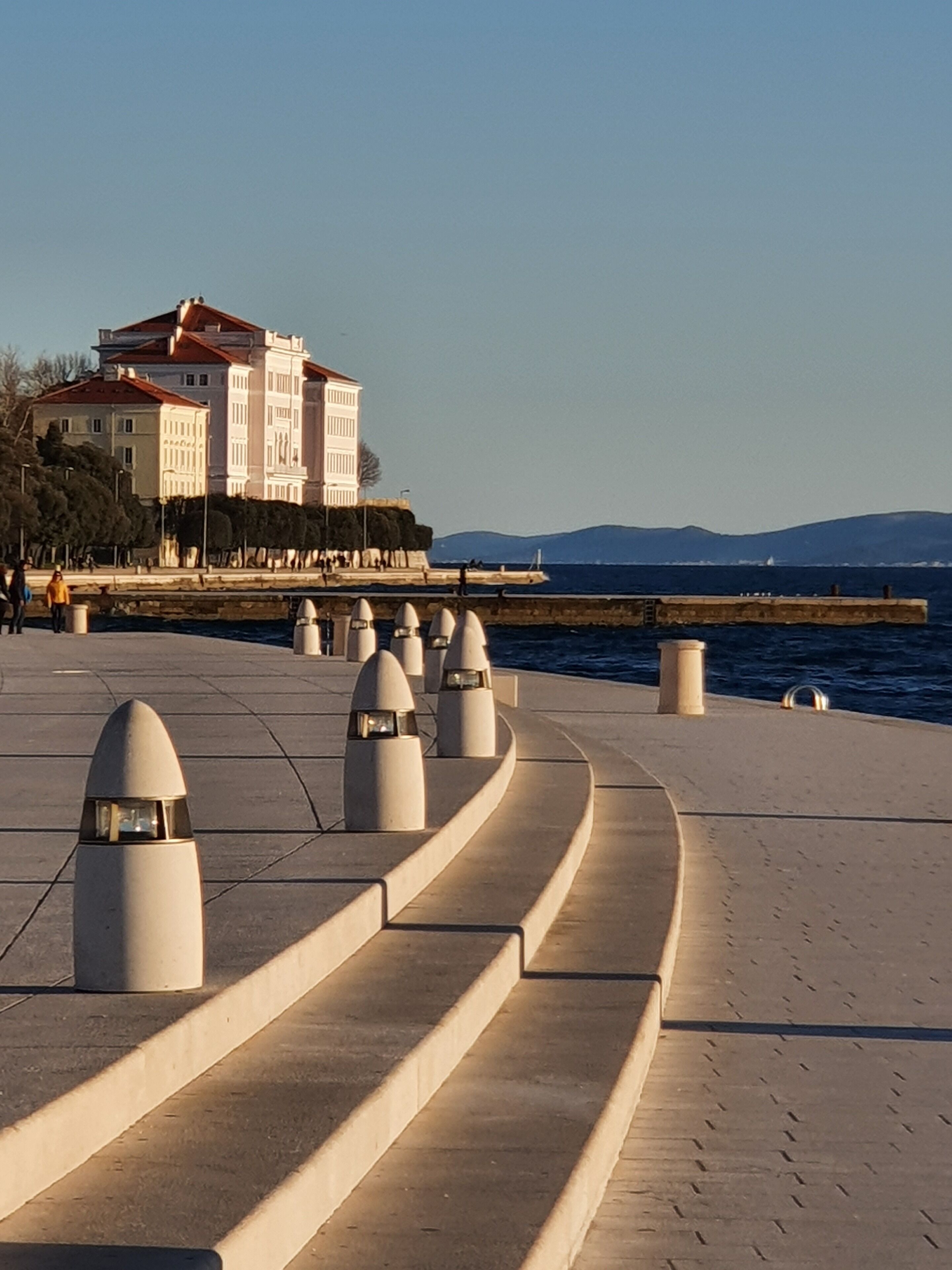 A waterfront promenade with a row of bollards and a large building with a red roof in the background.