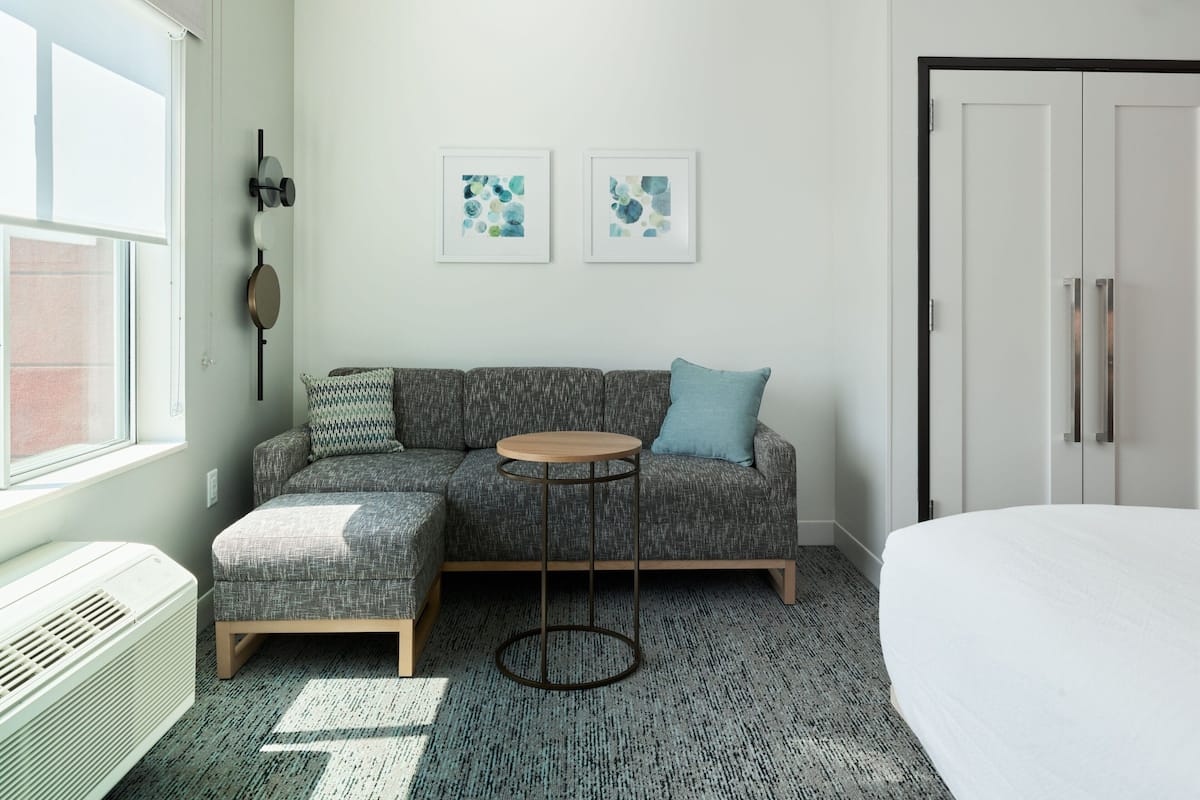 A modern room with a grey sectional sofa, a round wooden side table, a white radiator, and two abstract art pieces on the wall.