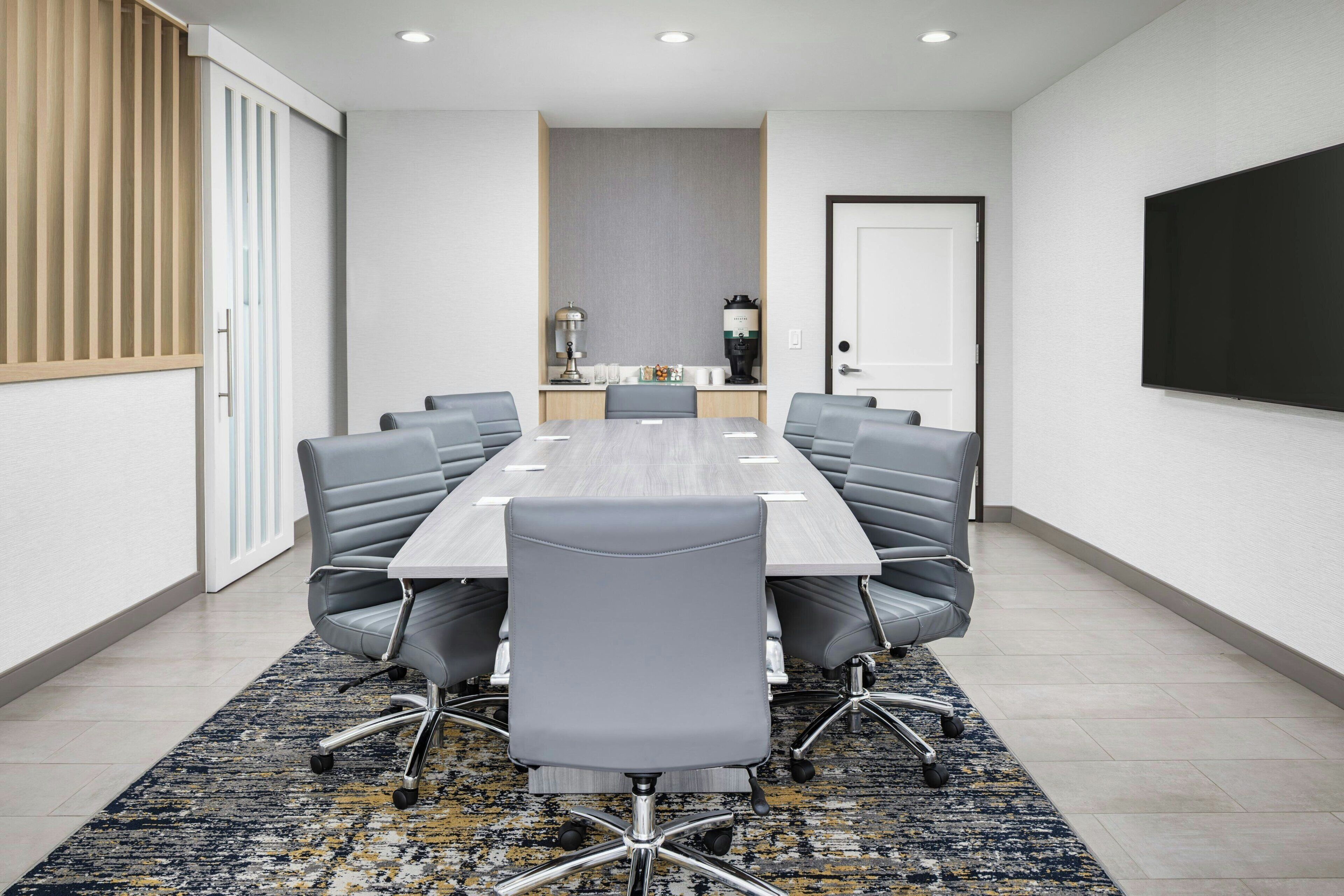 A conference room with a long table, grey chairs, a projector screen, and a coffee machine.