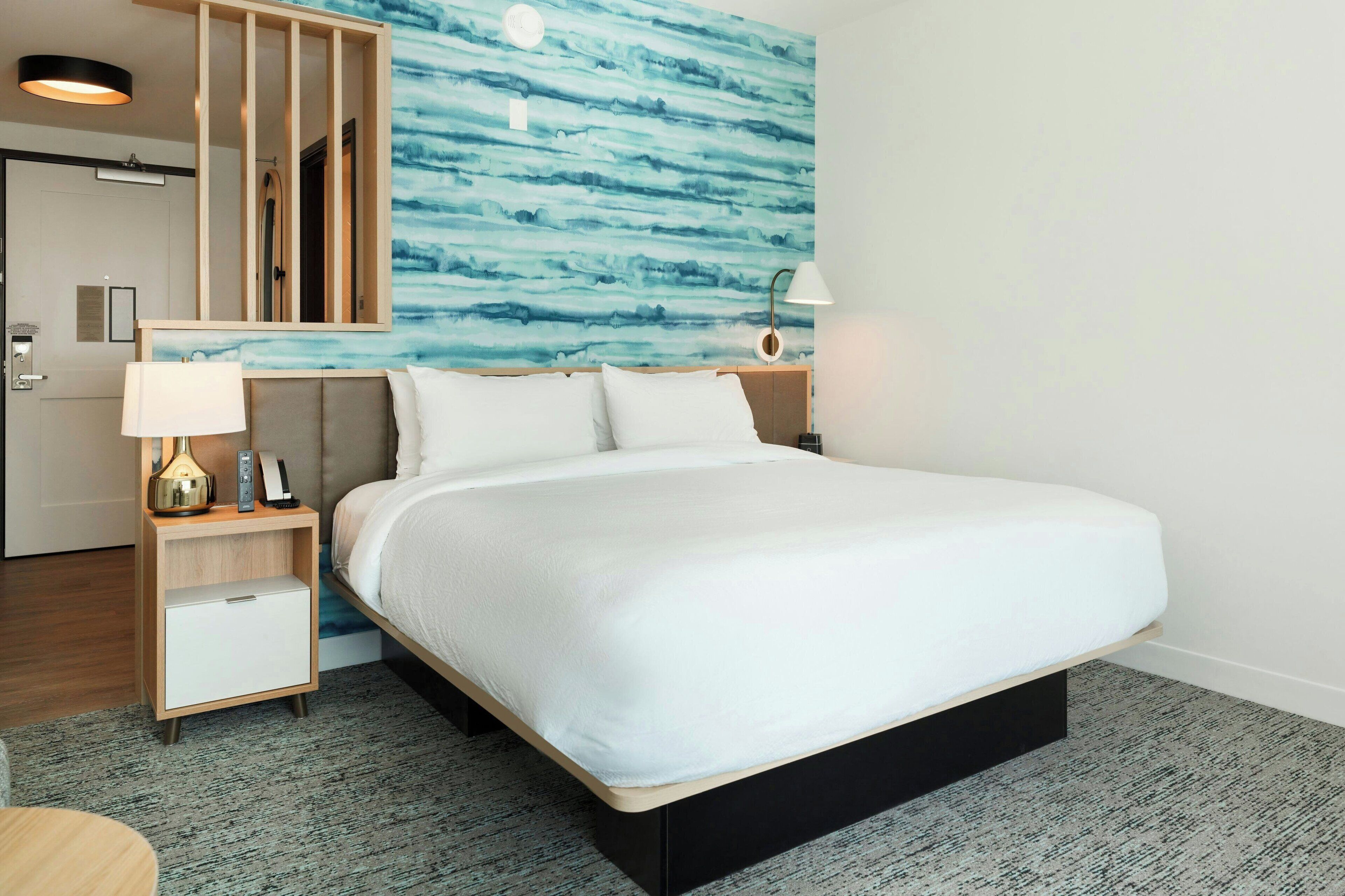 A hotel room with a bed, a nightstand with a lamp, a small cabinet, and a wall with a blue abstract design.