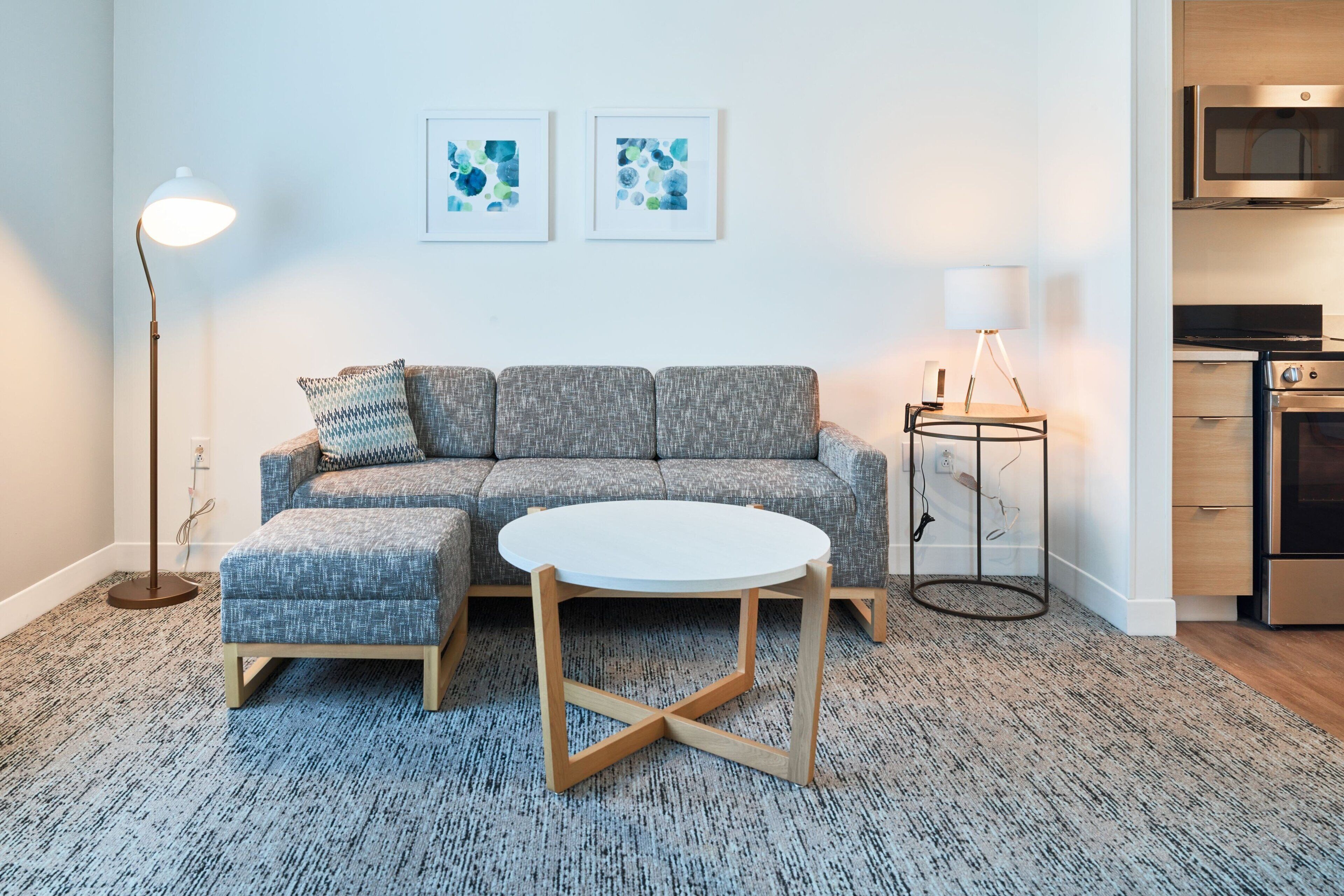 A modern living room with a sectional sofa, a round coffee table, a floor lamp, and a small side table with a lamp. There are abstract art pieces on the wall and a kitchen area with a microwave and oven.