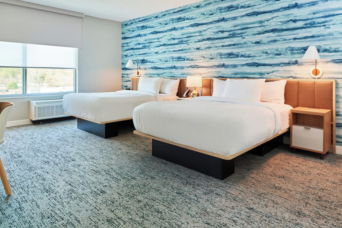 A hotel room with two beds, a large window, a wall mural, and a desk with a chair.