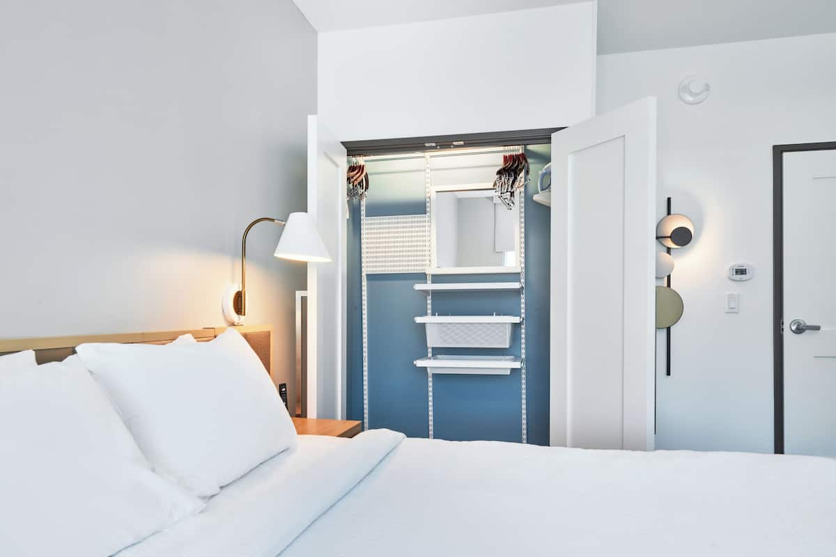A hotel room with a neatly made bed, a bedside lamp, a wardrobe with hanging items, and a door with a lock.