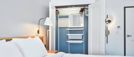 Habitación de hotel con una cama bien hecha, una lámpara de noche, un armario con ropa colgada y una puerta con cerradura.