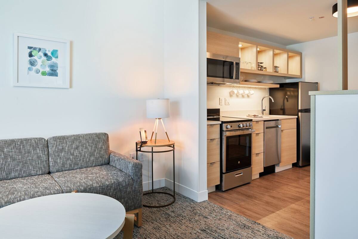 A modern kitchen with stainless steel appliances, a grey sofa, and a round table.
