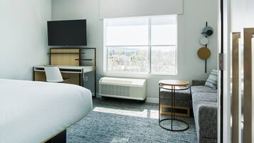 Quarto de hotel com cama, secretária com cadeira, televisão, radiador, sofá, mesa redonda pequena e janela com vista para edifícios.