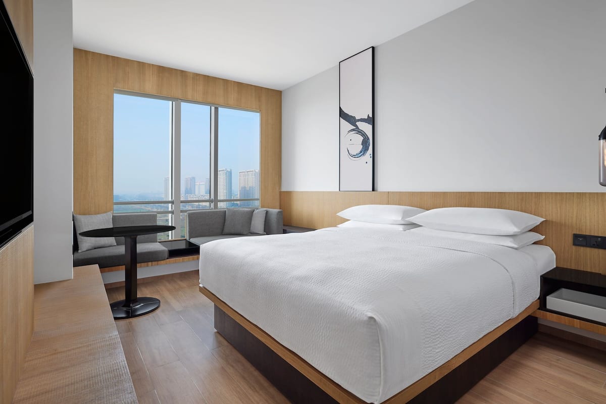 Una habitación de hotel moderna con una cama grande, una zona de estar con sofá y una pequeña mesa, y un ventanal con vistas a la ciudad.