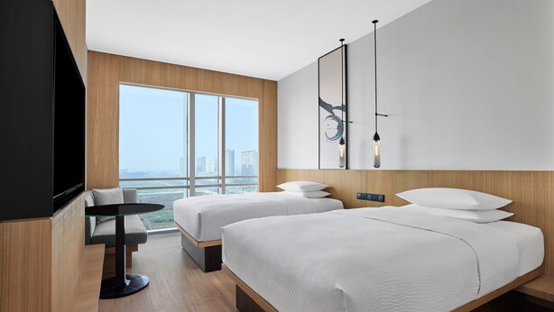 Habitación de hotel moderna con dos camas, un televisor, una mesita pequeña y un ventanal grande con vistas a la ciudad.