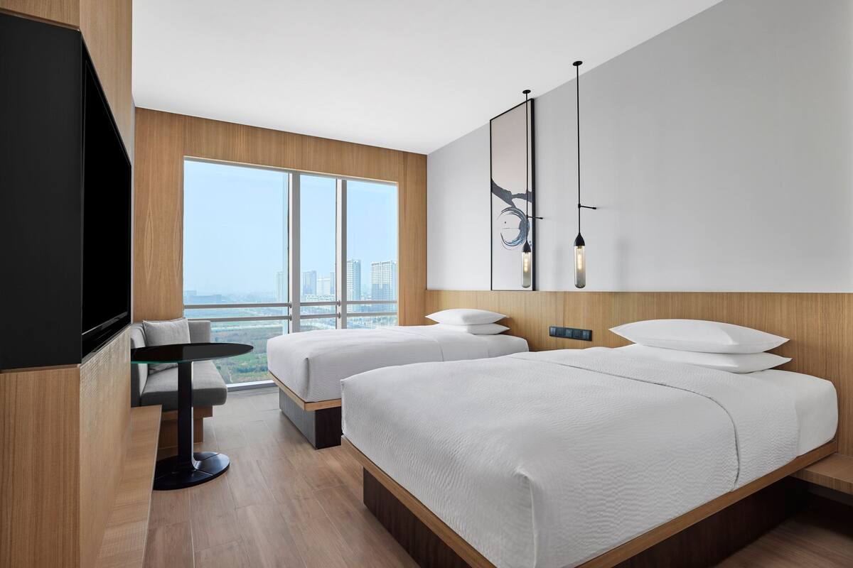 Habitación de hotel moderna con dos camas, un televisor, una mesita pequeña y un ventanal grande con vistas a la ciudad.