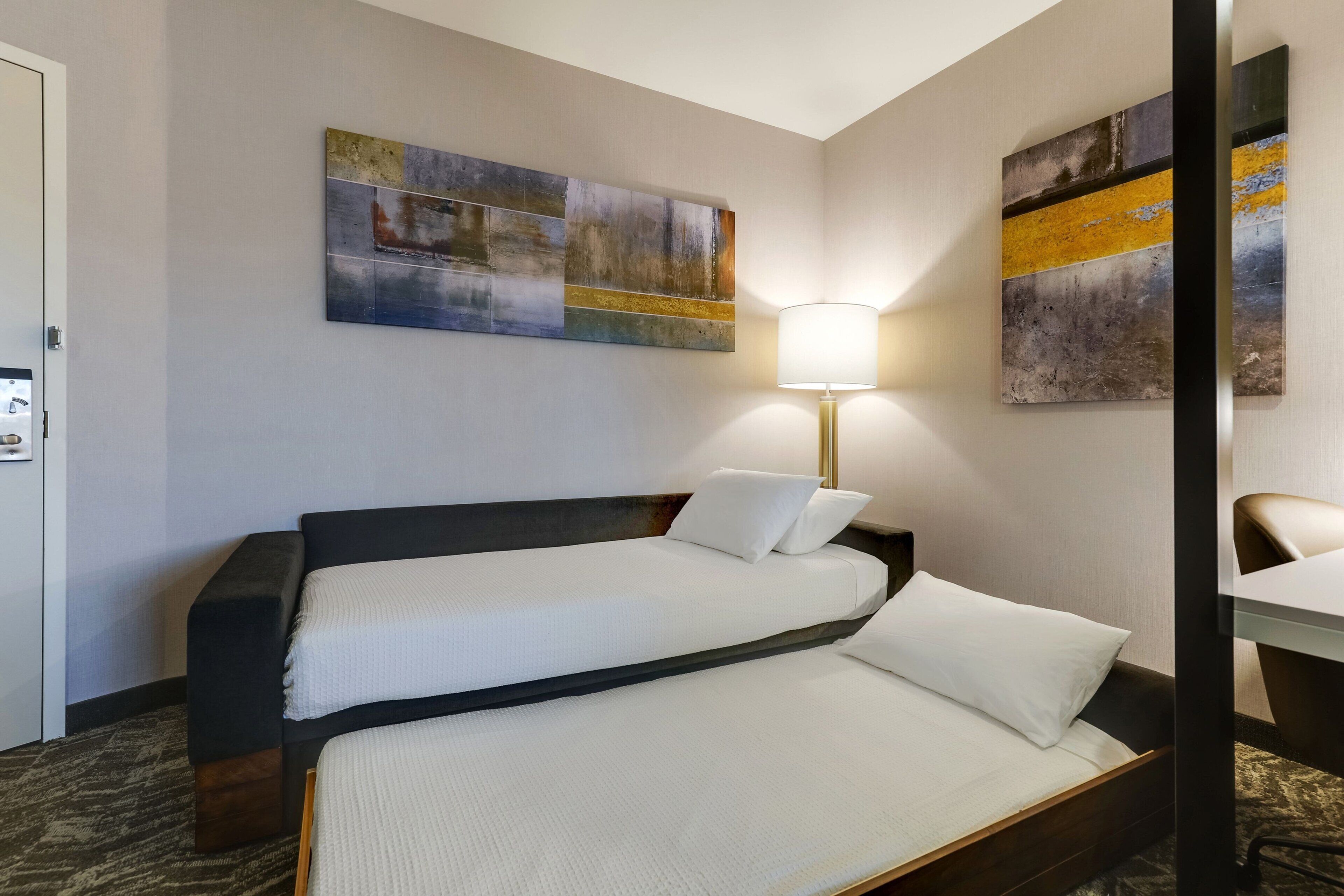 A hotel room with a double bed, a bedside table, a lamp, and a large abstract painting on the wall.