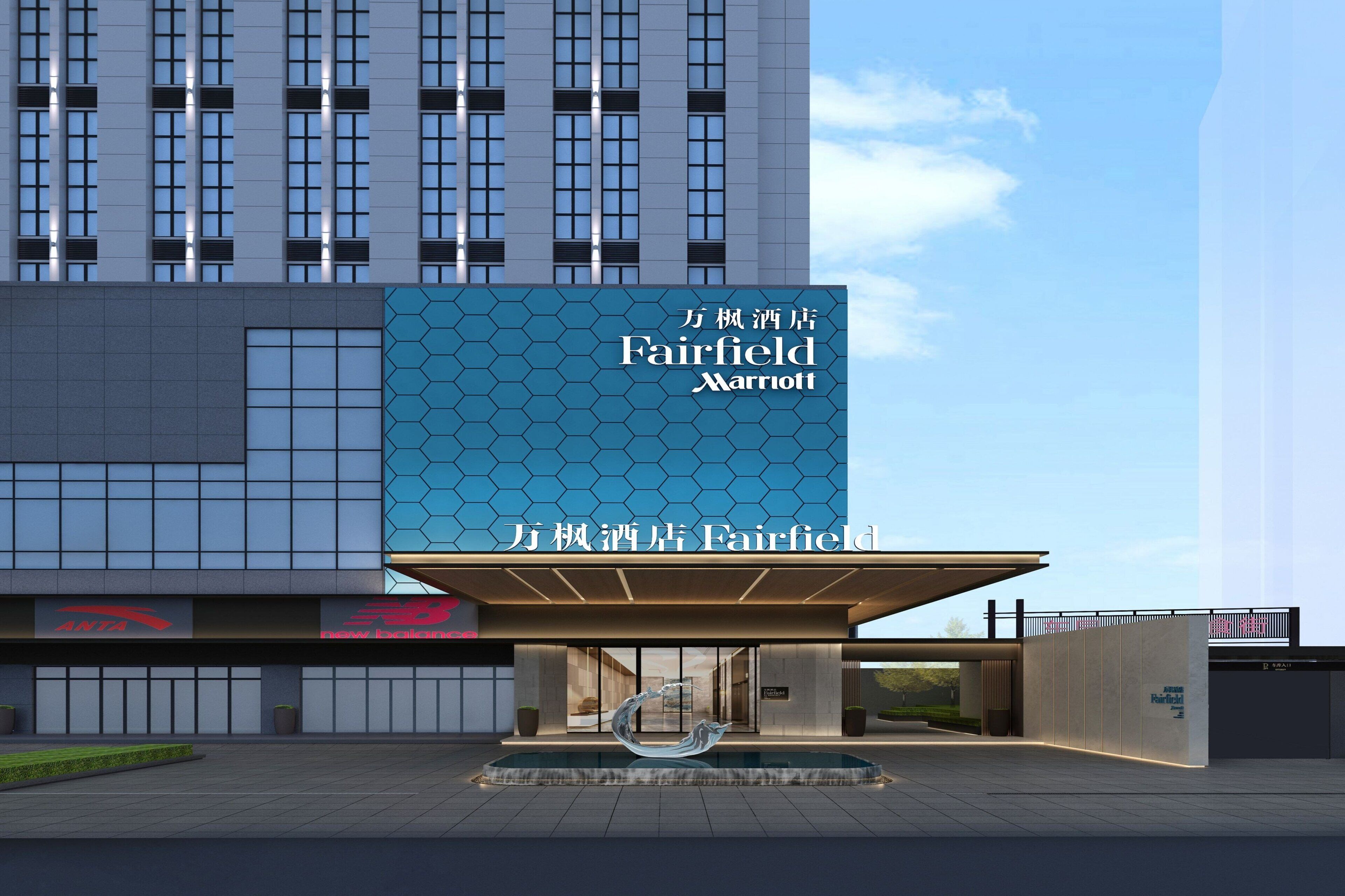 Una entrada de hotel moderna con un gran letrero de Fairfield Marriott, fachada de vidrio geométrica y una fuente en primer plano.