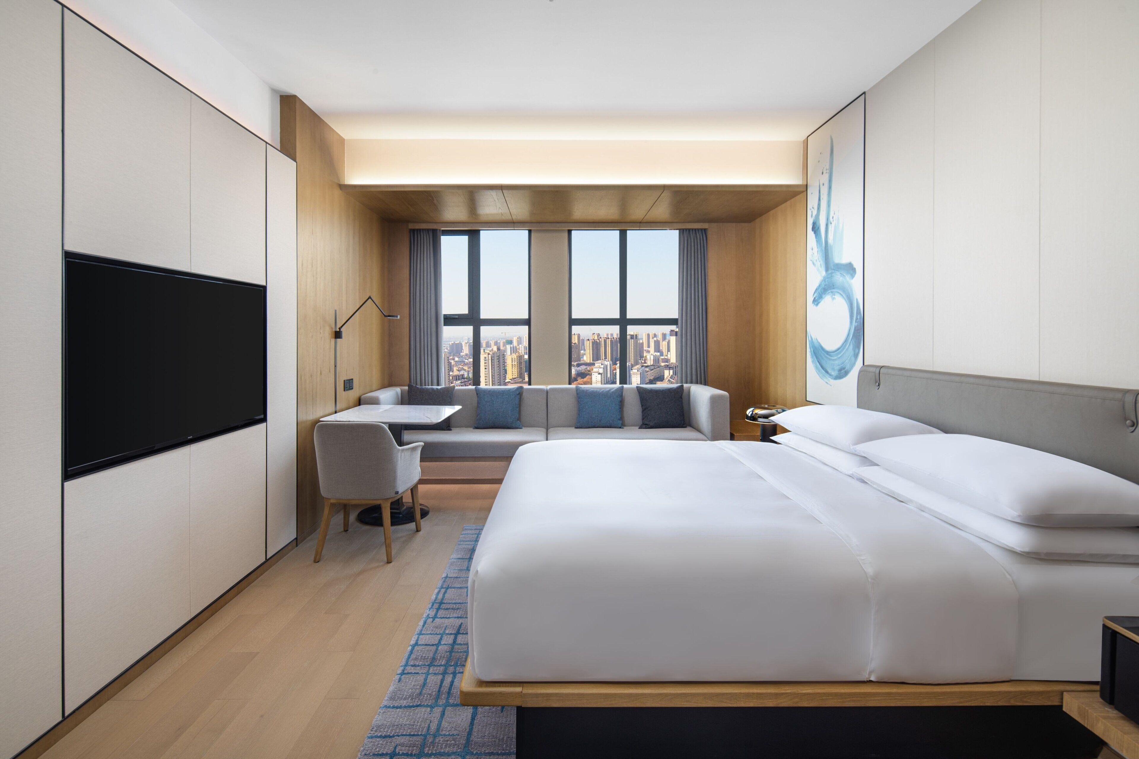 Quarto de hotel moderno com uma cama grande, uma escrivaninha e um sofá. Vê-se uma vista da cidade pelas janelas.