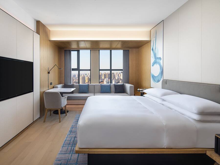 A modern hotel room with a large bed, a desk, and a sofa. There is a city view through the windows.