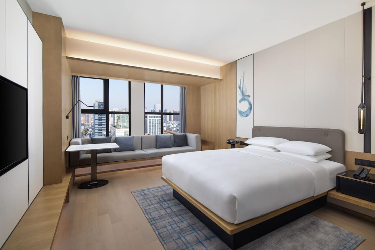Una habitación de hotel moderna con una cama grande, una zona de estar con sofá y mesa, un televisor y vistas a la ciudad a través de amplias ventanas.