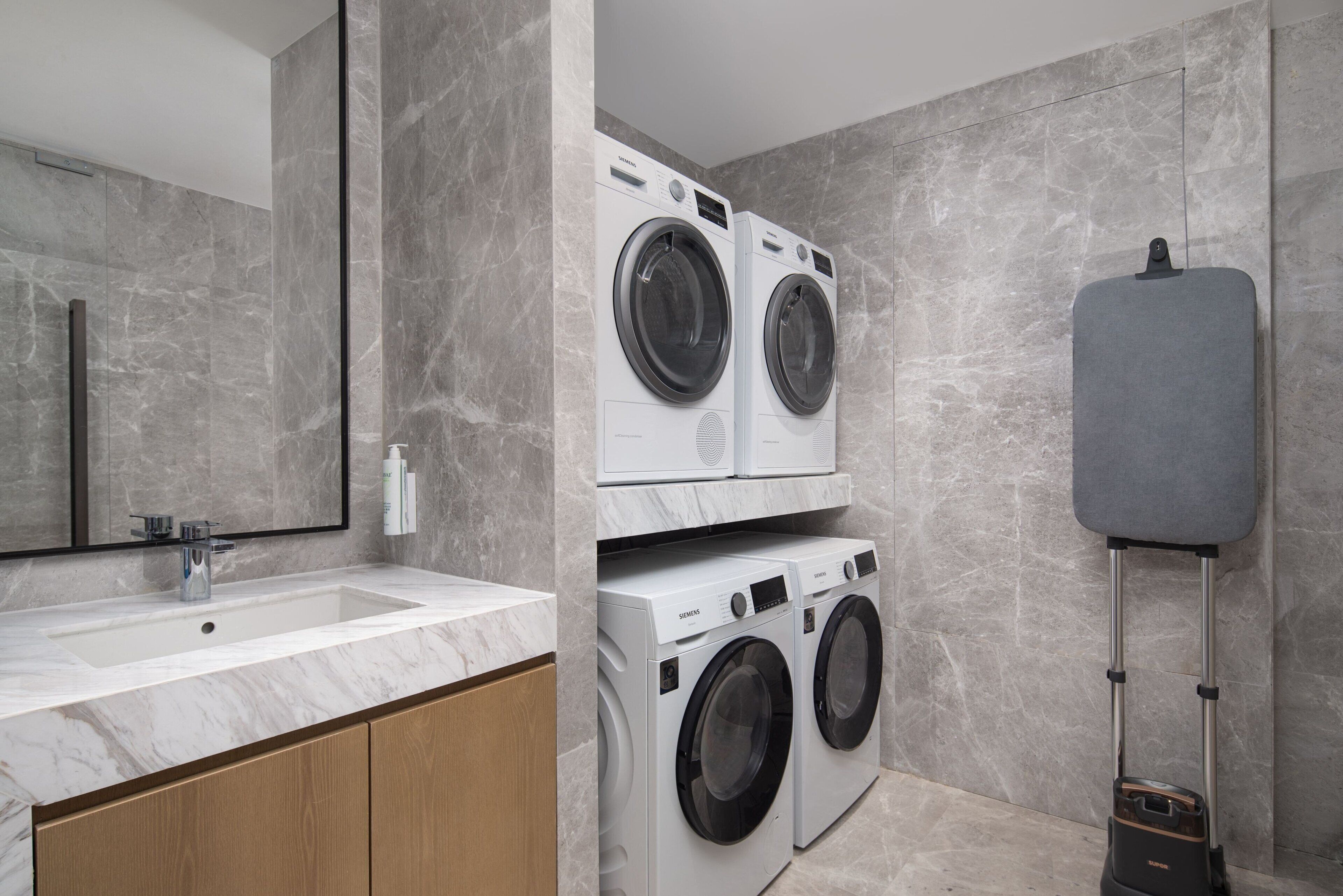 A modern bathroom with a marble sink, two stacked front-loading washing machines, and a grey portable toilet.