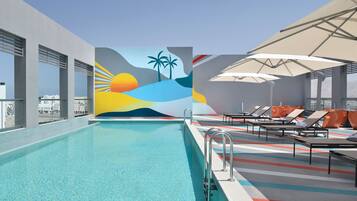 A rooftop pool area with a mural of a sunset and palm trees, lounge chairs, and umbrellas.