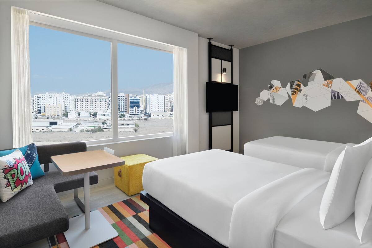 Habitación de hotel con un ventanal grande, una cama, un sofá, una mesita y un tapete colorido.