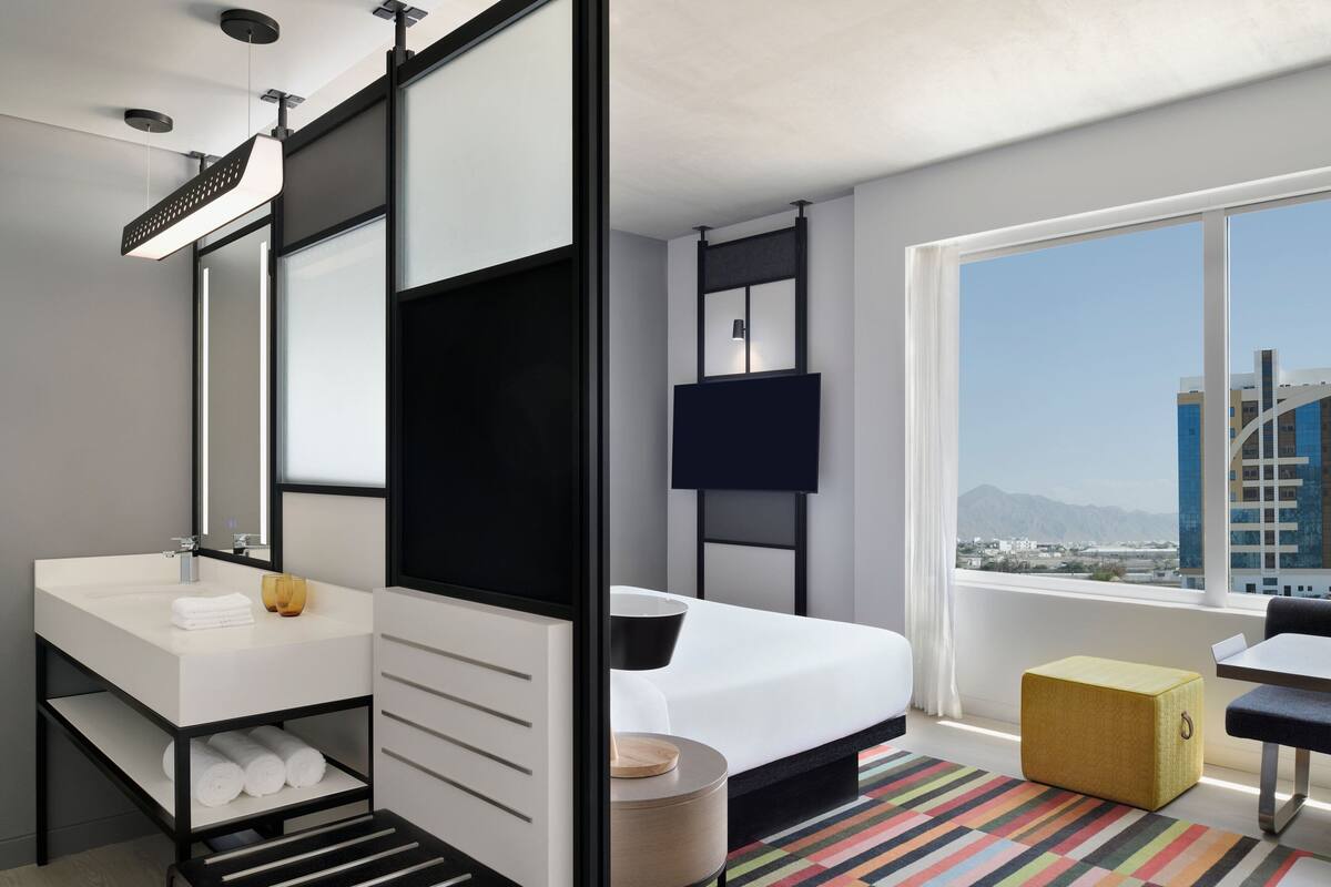 Una habitación de hotel moderna con una cama grande, un lavamanos blanco, un espejo, un televisor de pantalla plana y un ventanal que ofrece vistas a la ciudad.