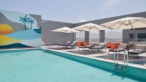 A swimming pool with lounge chairs and umbrellas, a mural with palm trees and a sunset, and a view of distant mountains.