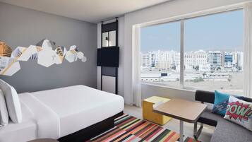 Una habitación de hotel moderna con una cama grande, una mesita pequeña, un sofá, una alfombra colorida a rayas y vistas a la ciudad a través de un ventanal amplio.