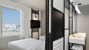 A modern hotel room with a large bed, a bathroom with a white sink and mirror, and a city view through the window. - Aloft by Marriott Muscat (Muscat)