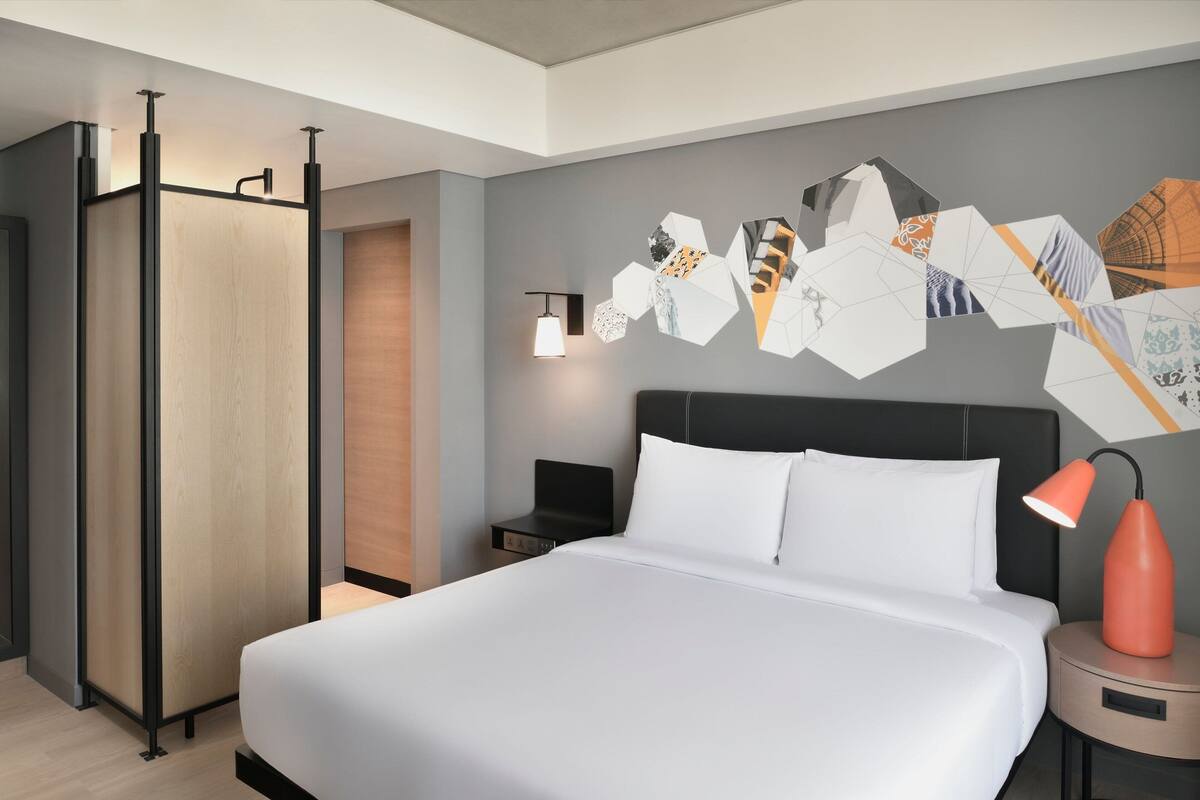 Habitación de hotel moderna con una cama grande, una mesita de noche, una lámpara de pared y una obra de arte geométrica en la pared.