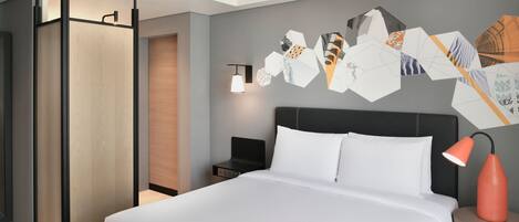 Habitación de hotel moderna con una cama grande, una mesita de noche, una lámpara de pared y una obra de arte geométrica en la pared.