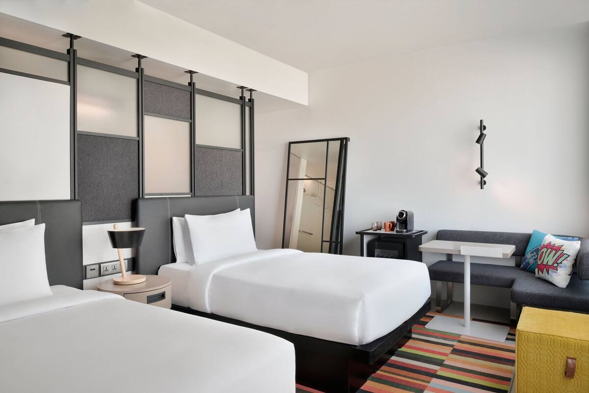 Habitación de hotel con dos camas, un escritorio, un sofá, un espejo y una mesita de noche con lámpara.