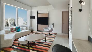 A modern living room with a sofa, a round coffee table, a striped rug, a white chair, and a city view through large windows.