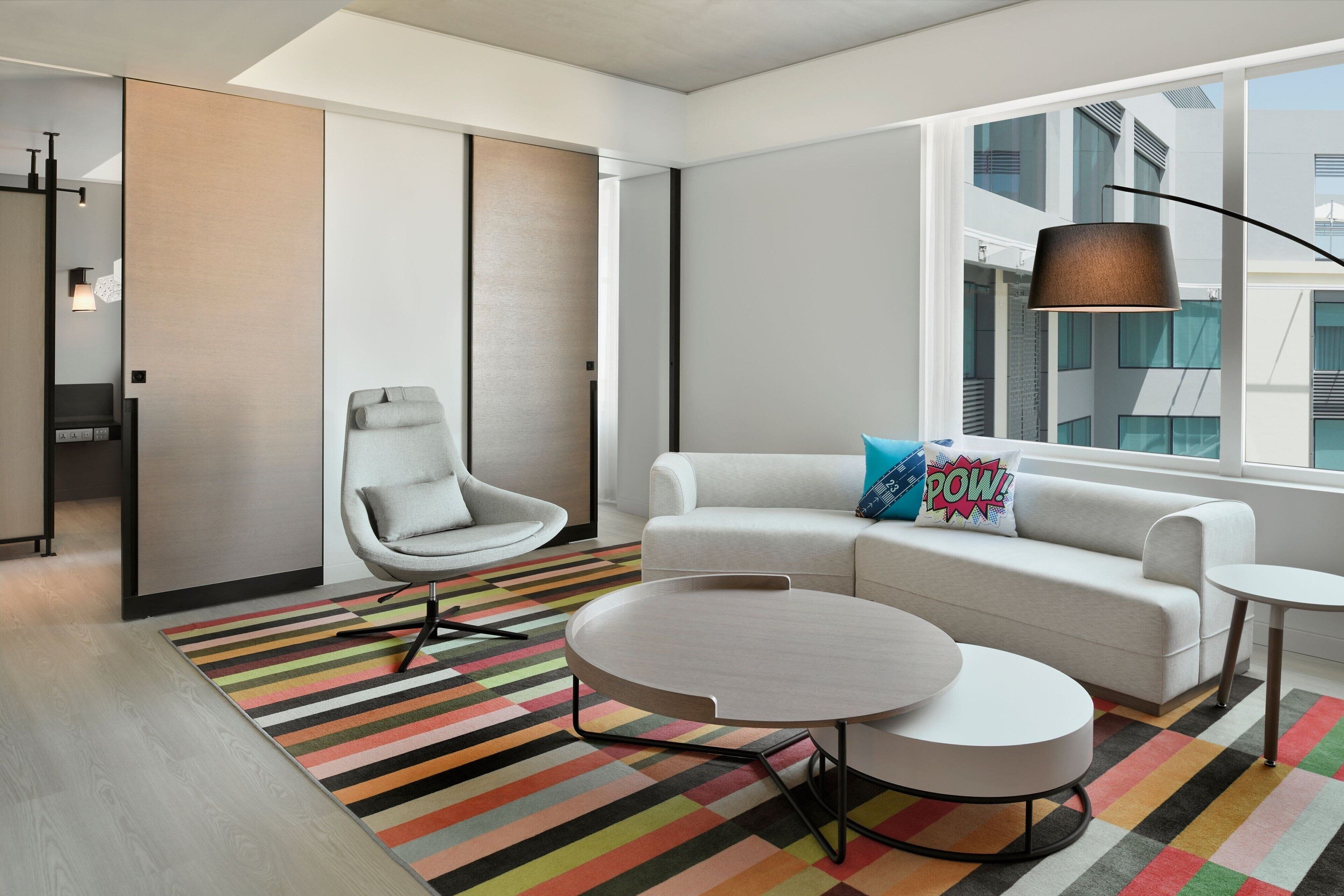A modern living room with a white sofa, a round coffee table, a striped rug, and a large window with a view of buildings.