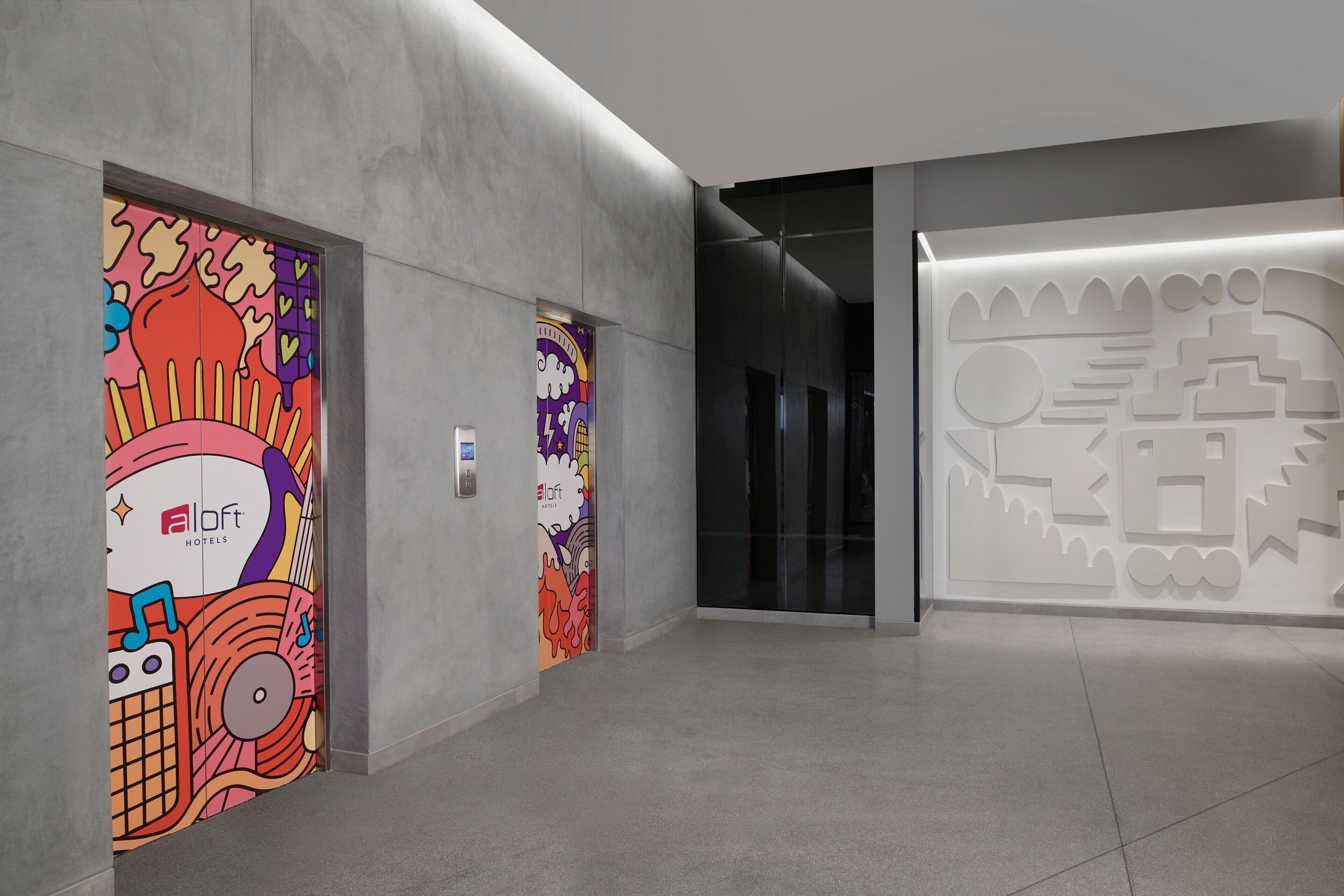 Un hall d’hôtel moderne au design minimaliste, agrémenté d’une fresque murale lumineuse et colorée sur le mur de gauche et d’une œuvre d’art géométrique sur le mur de droite.