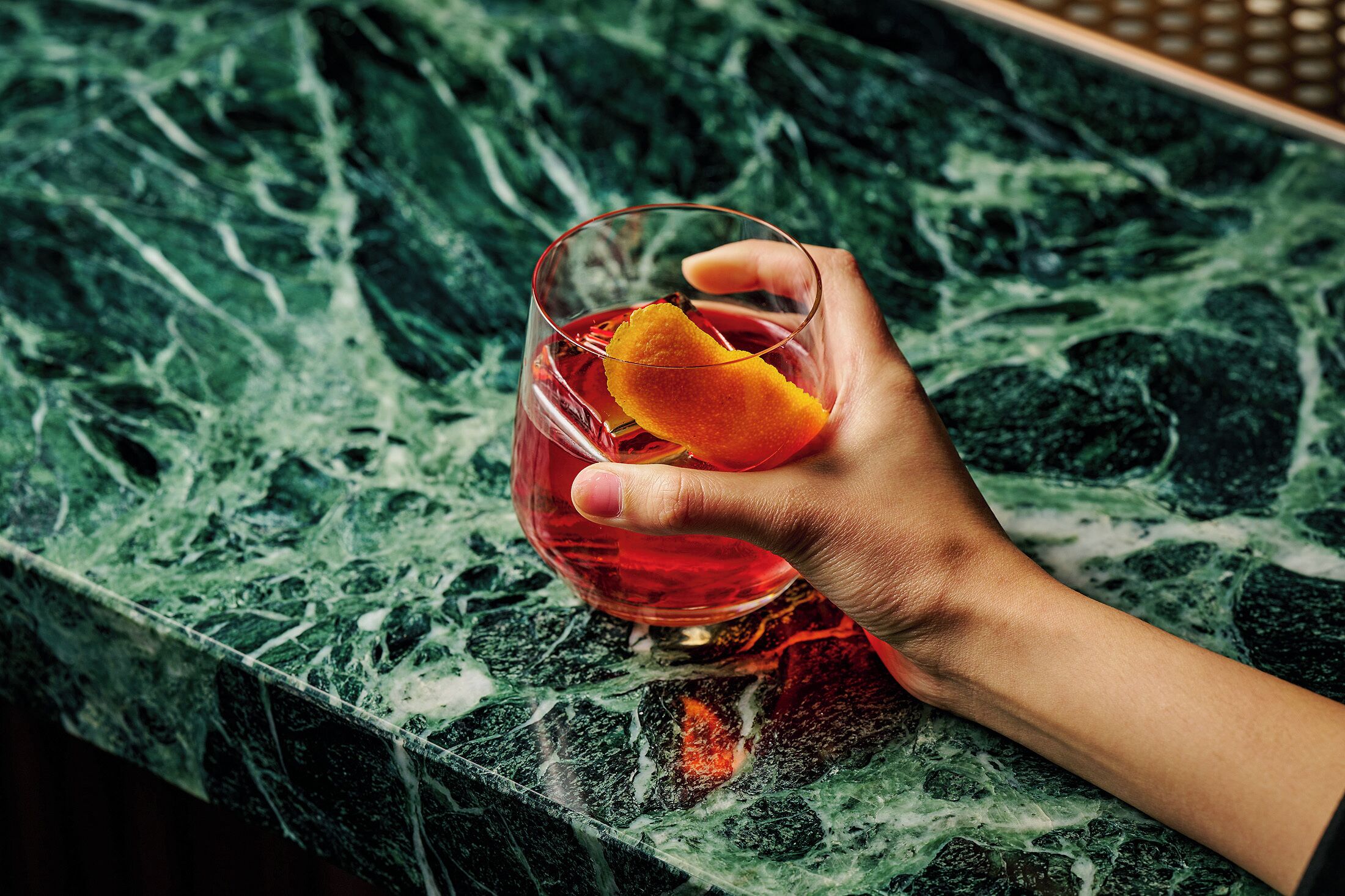 Una mano sostiene un vaso con un líquido rojo y una rodaja de naranja sobre una superficie de mármol.