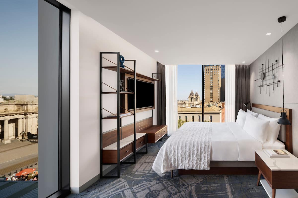 A modern hotel room with a large bed, a TV, a desk, and a view of a cityscape.