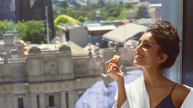 A person in a white robe sitting by a window overlooking a cityscape with buildings and a table with various dishes. - Le Méridien Melbourne (Melbourne)