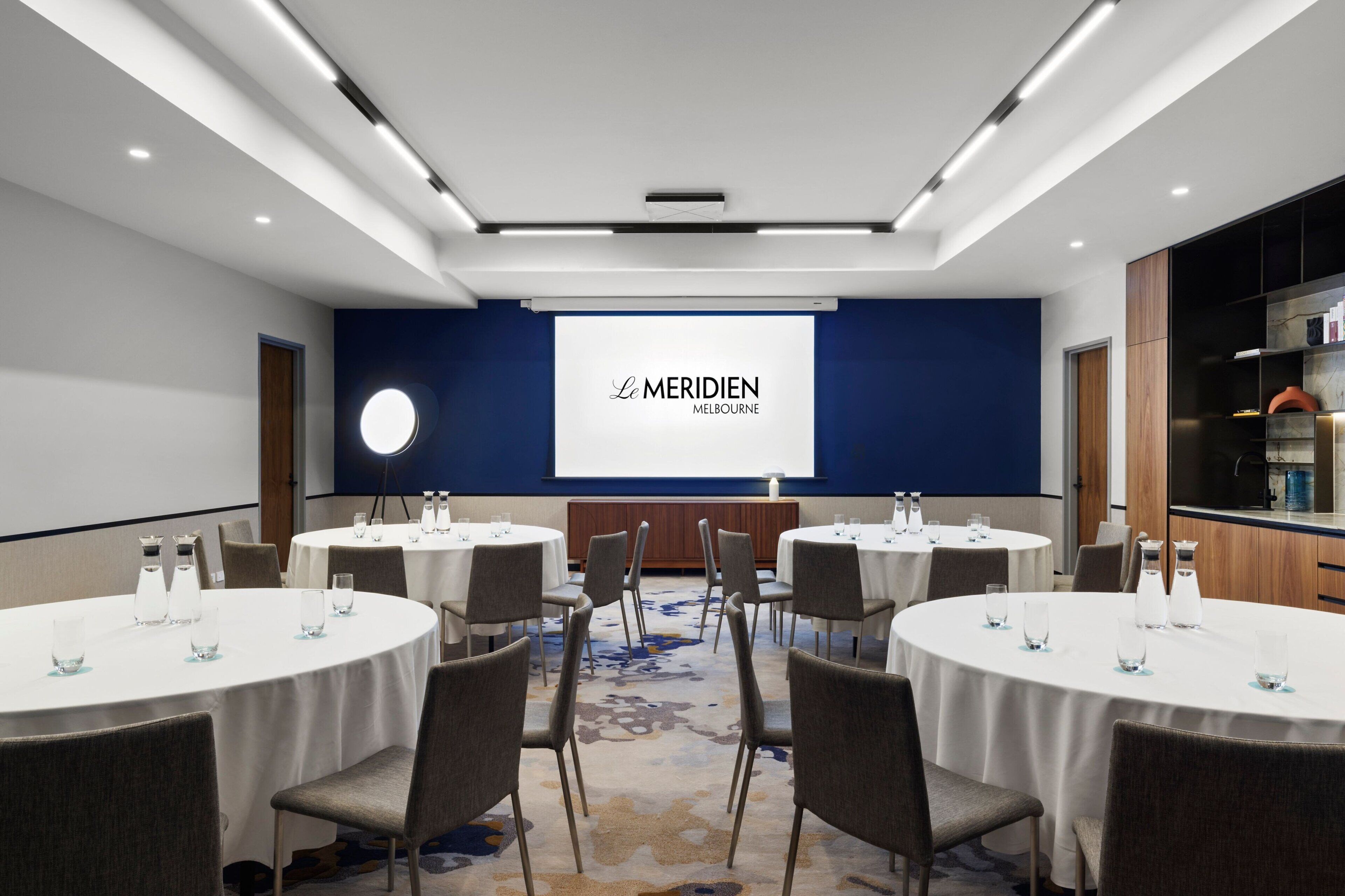 Une salle de conférence équipée de plusieurs tables rondes, de chaises et d’un écran de projection affichant le nom « Le Meridien Melbourne ».