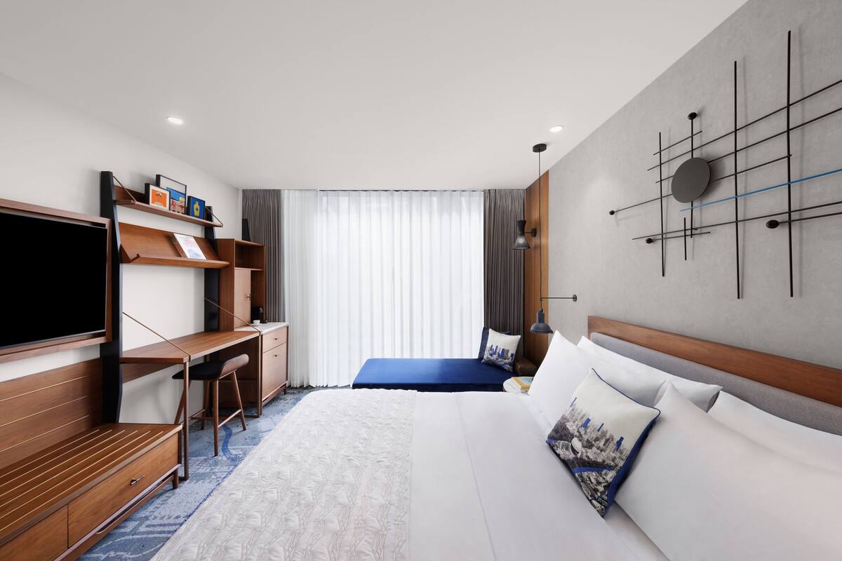 A modern hotel room with a bed, a desk, a TV, and a wall-mounted artwork.