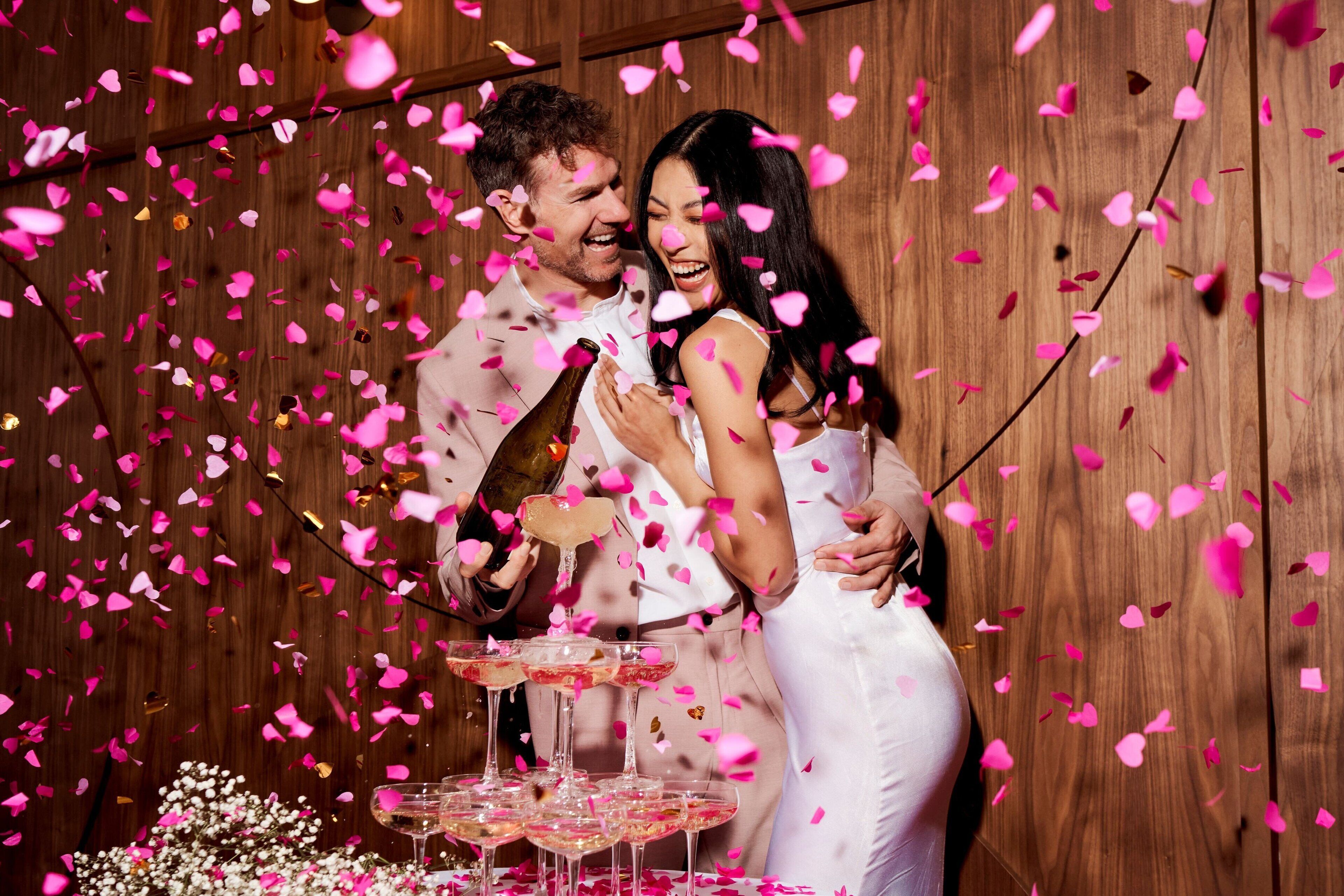 Um casal comemorando com champanhe e pétalas de rosas em frente a um painel de madeira.