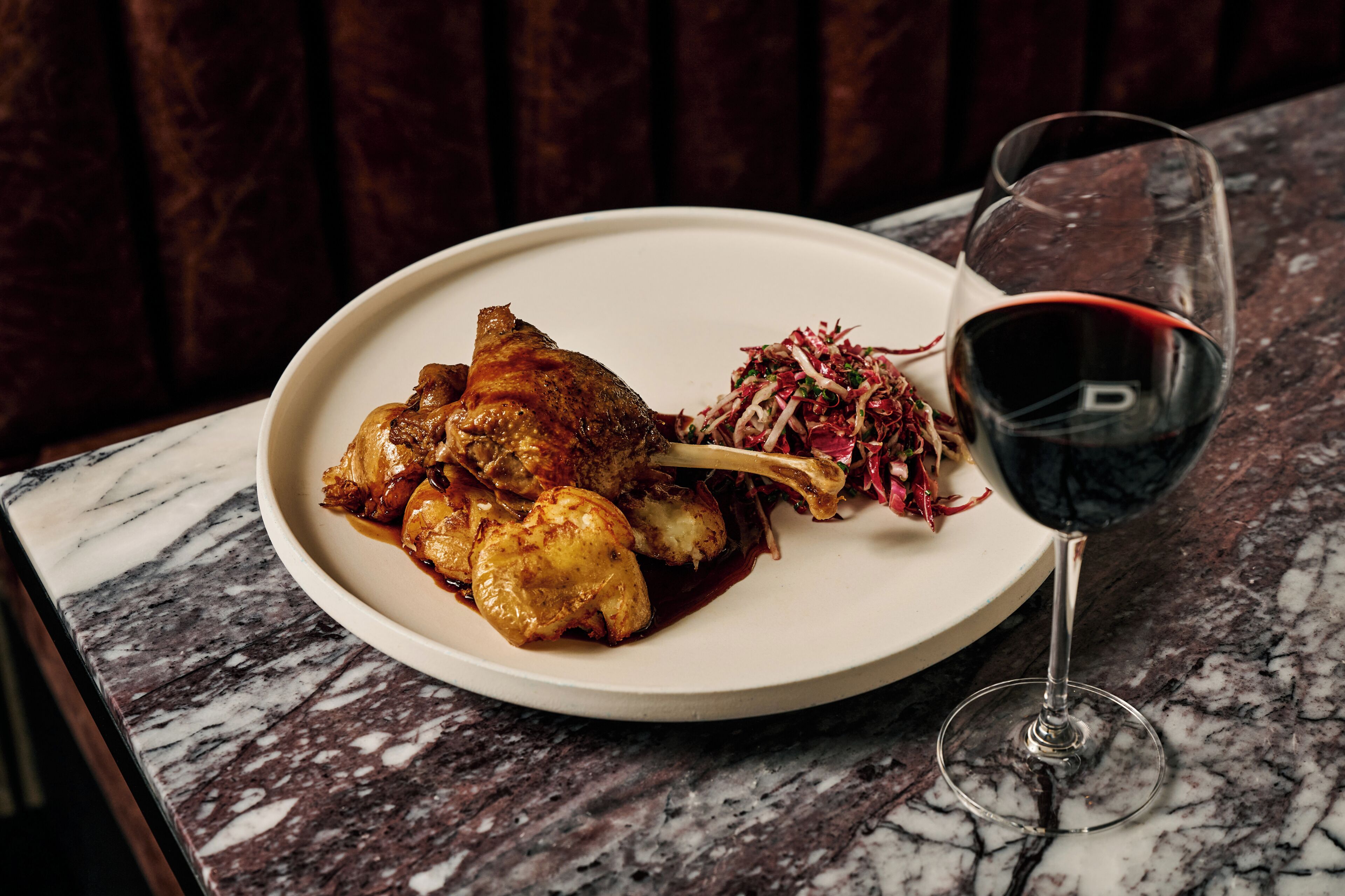 A plate of roasted chicken with a side salad and a glass of red wine on a marble table.