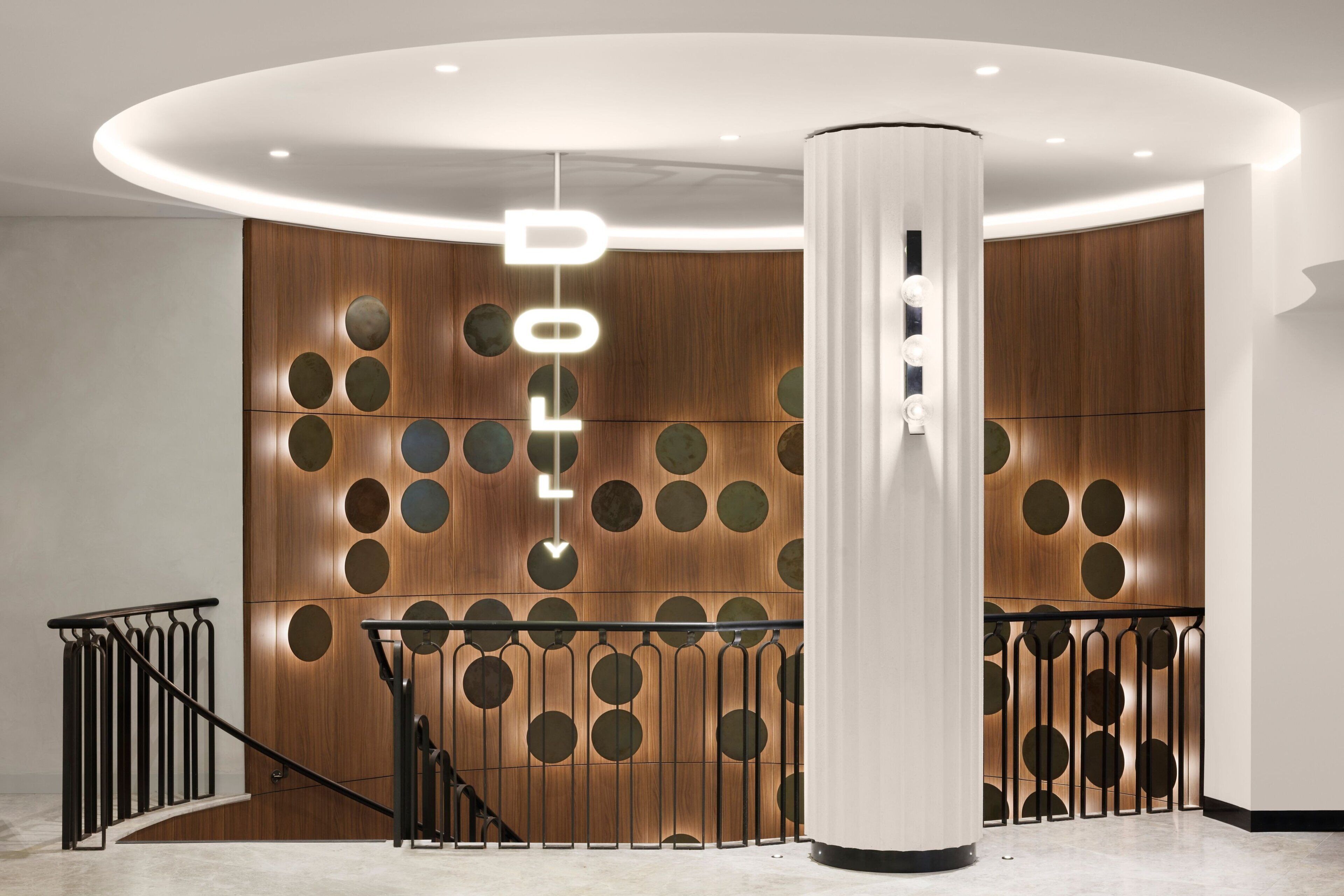 A modern lobby with a wooden wall featuring circular cutouts, a central column, and a staircase with metal railings.
