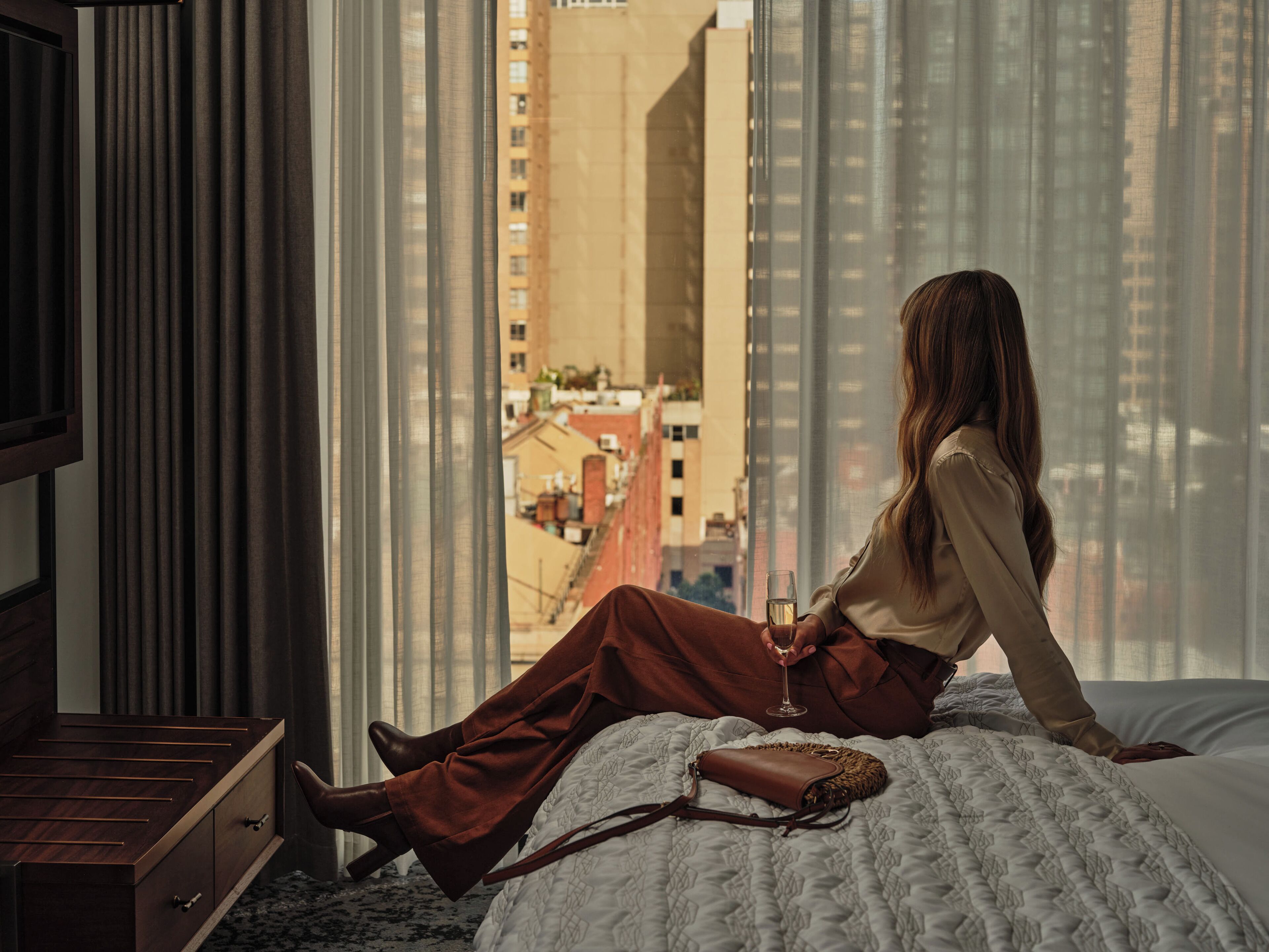 A person sitting on a bed with a view of a cityscape through large windows.