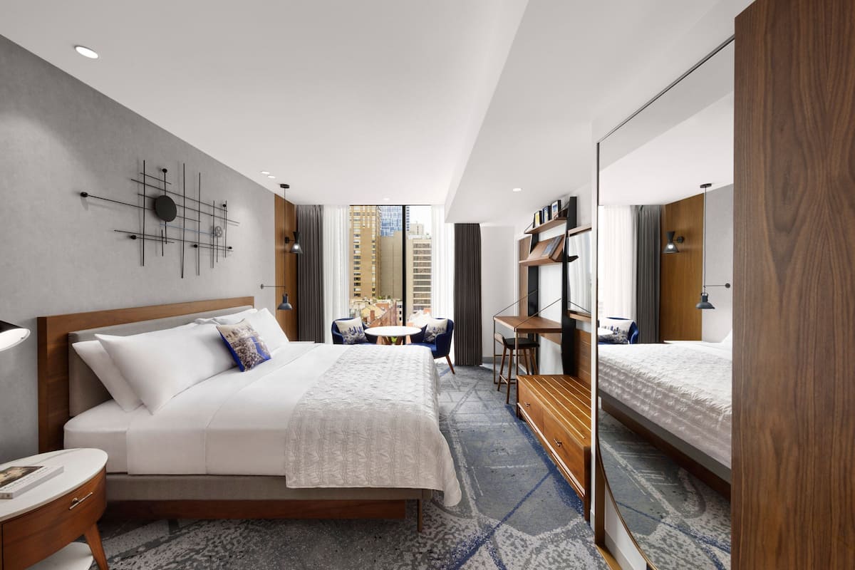 A modern hotel room with a large bed, a desk, a chair, a small table, and a large mirror.
