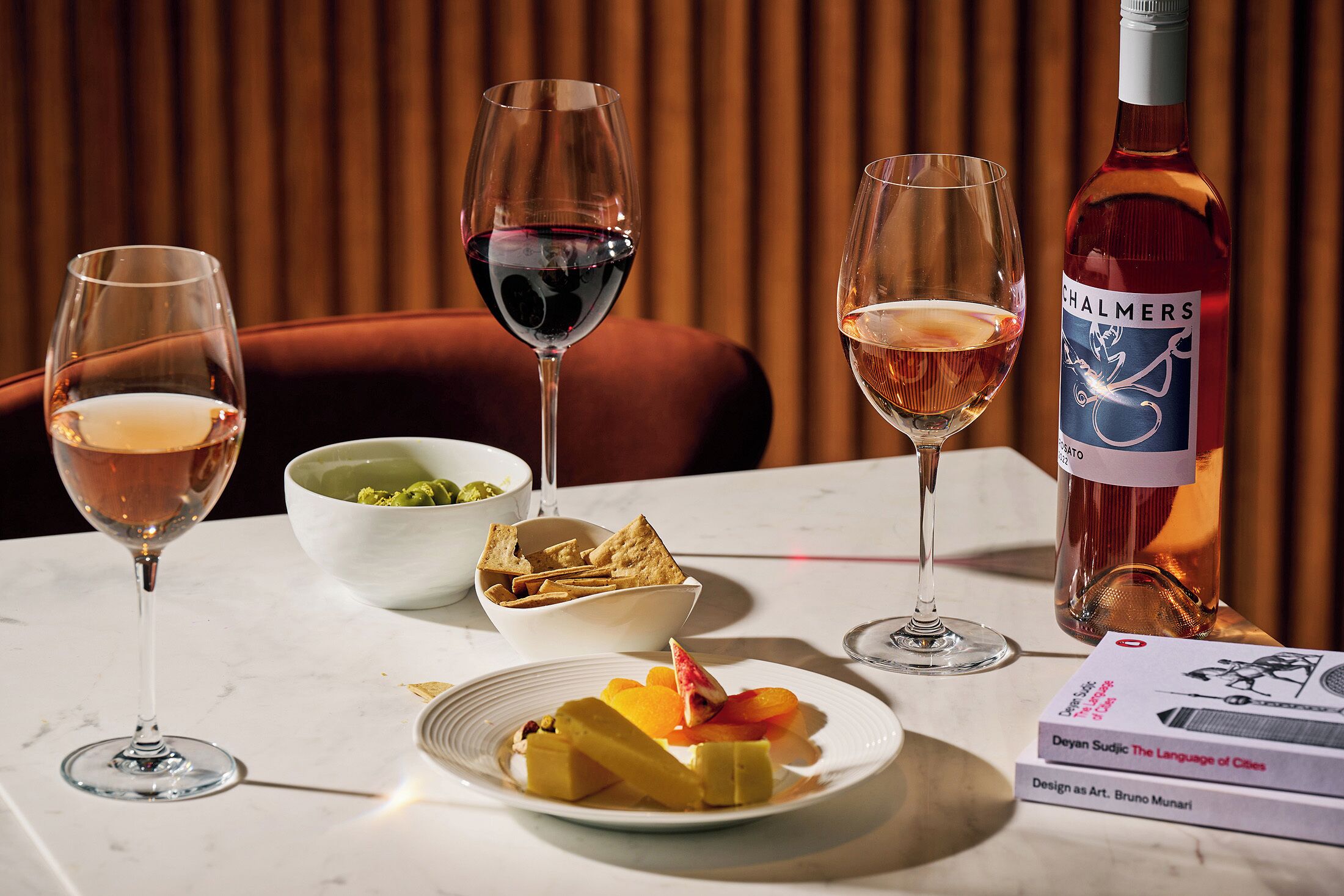 A table with wine glasses, a bottle of wine, a bowl of snacks, a plate of fruit, and a book.