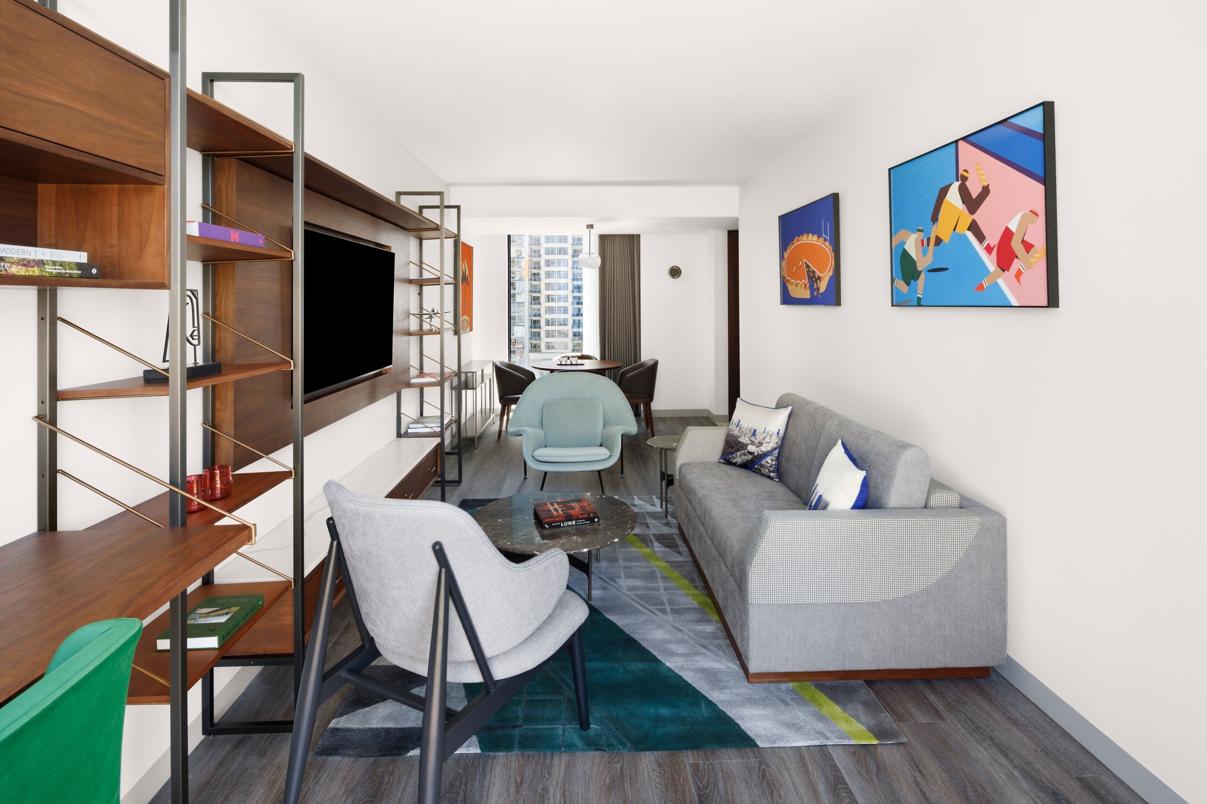 Una sala moderna con un sofá gris, una mesa de centro y un televisor sobre una estantería de madera. Hay dos sillones, una mesa de comedor y obras de arte en las paredes.